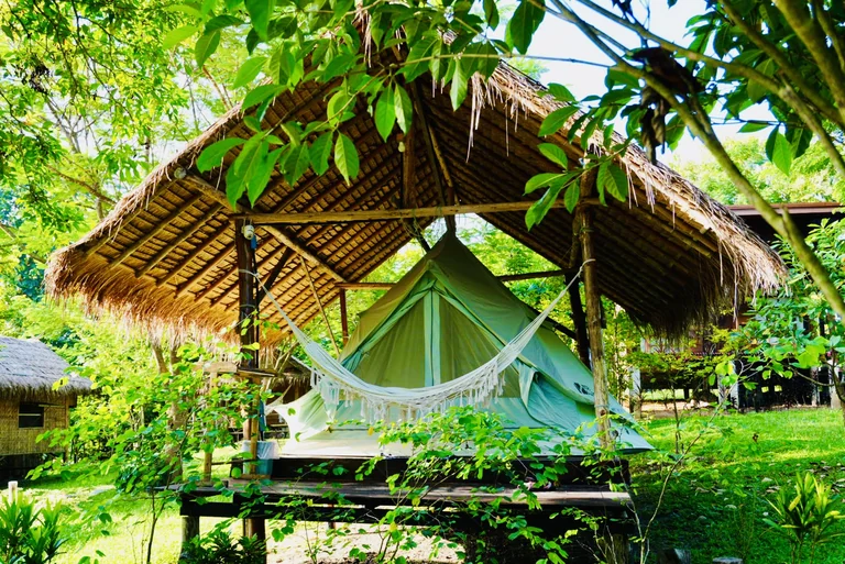 A raised canvas tent and hammock beneath a thatched shelter in the garden