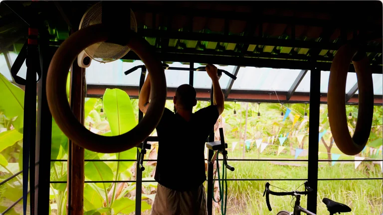 A guest using the pull-up bar, with gymnastic rings and banana plants around the gym