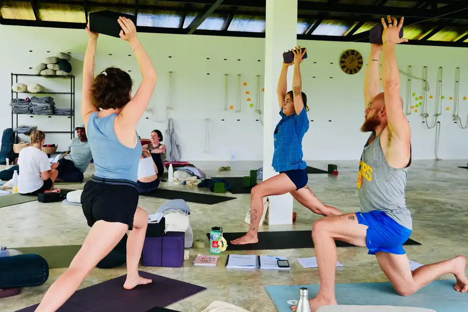 Teacher training students in a lunge with blocks held overhead in the open-air shala, September 2026